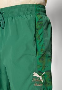 Pantalones cortos verdes con un panel lateral de camuflaje, cintura elástica con cordón y logotipo bordado de KidSuper Studios con la silueta de Puma.