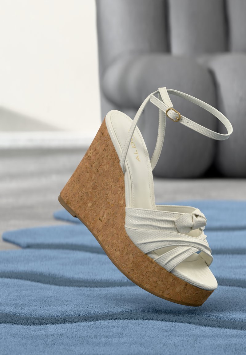 Sandale compensée blanche avec plateforme en liège, bride à la cheville et détail noué à l’avant reposant sur un tapis bleu texturé.