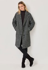 Cappotto grigio a scacchi con una vestibilità rilassata, ampi revers e chiusura a bottone. Abbinato a una maglietta scura e pantaloni neri, con stivali alla caviglia.