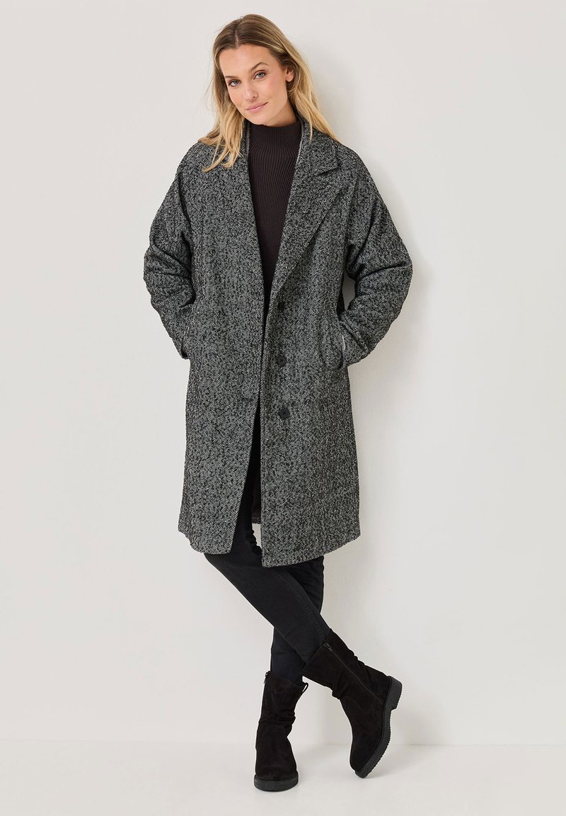 Cappotto grigio a scacchi con una vestibilità rilassata, ampi revers e chiusura a bottone. Abbinato a una maglietta scura e pantaloni neri, con stivali alla caviglia.