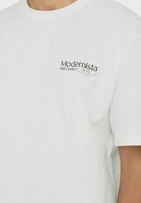 T-shirt blanc en coton avec un petit logo en texte noir et rose "Modernista Mid Century NYC" sur le côté gauche de la poitrine. Manches courtes, coupe décontractée.