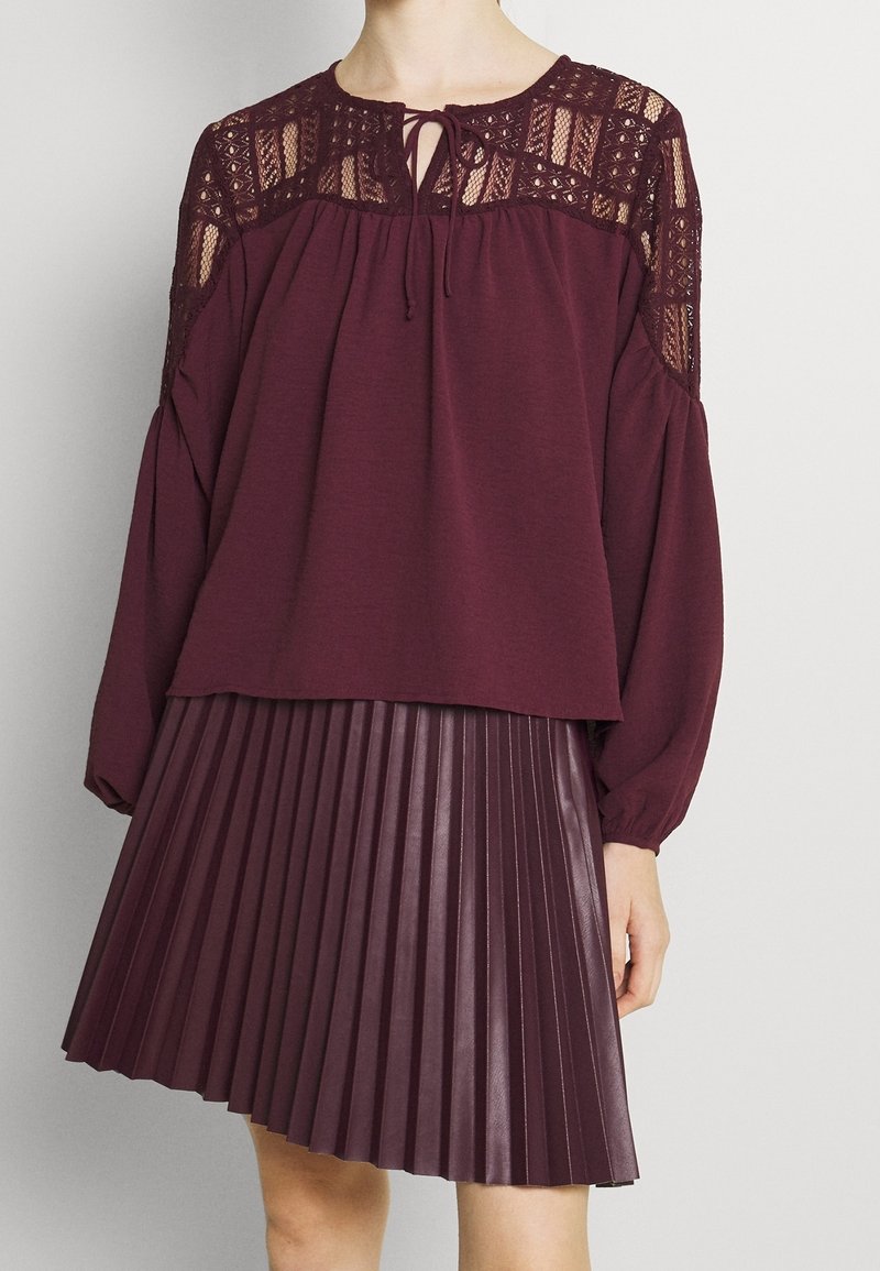 Bordeaux bluse med blonde detaljer og en bindebøjle, parret med en plisseret bordeaux nederdel. Begge har en blød tekstur og en løs pasform.