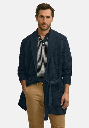Man draagt een marineblauwe gebreide cardigan met patroon en ceintuur over een gestreept poloshirt en beige broek, staand met één hand in de zak.
