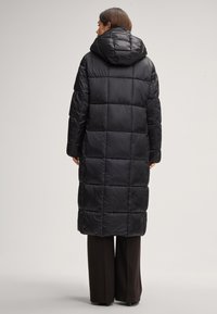 Cappotto lungo, imbottito e nero con cappuccio e motivo trapuntato. Caratterizzato da una texture lucida e liscia e una vestibilità comoda, indossato sopra pantaloni scuri.