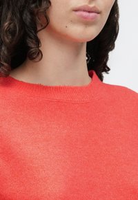 Rød strikket genser med rund hals, preget av en myk tekstur og jevn søm, som fremhever det glatte stoffet og den livlige fargen.