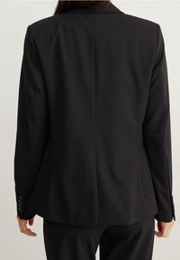 Personne portant un blazer noir ajusté et un pantalon noir assorti, vue de dos avec les bras détendus le long du corps.