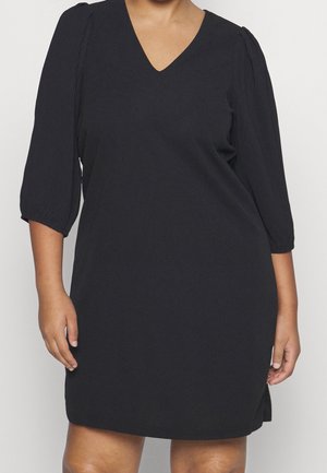 Mujer con un vestido negro hasta la rodilla con mangas abullonadas hasta tres cuartos y escote en V, de pie frente a un fondo liso.