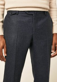 Texturované tmavě modré kalhoty s dvěma knoflíky, plochým předním dílem, bočními kapsami a upraveným střihem, zobrazené se rukama ležícími na boky.