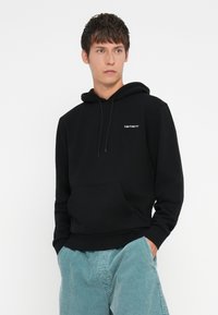 Black hoodie with a front pocket, drawstring hood, and white logo on the left chest. Model wears light blue corduroy pants.