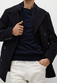 Cappotto navy doppiopetto con ampi risvolti e bottoni. Indossato sopra un maglione navy abbinato, accompagnato da pantaloni chiari.