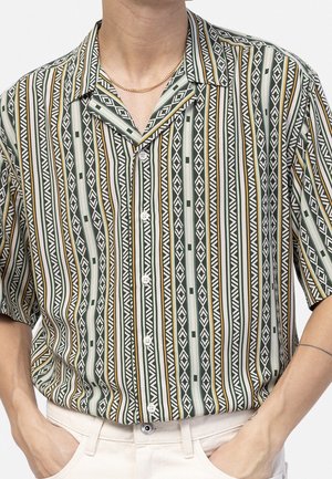 Homme portant une chemise à manches courtes boutonnée avec des rayures géométriques verticales vertes, blanches et moutarde, rentrée dans un pantalon blanc avec les mains dans les poches.