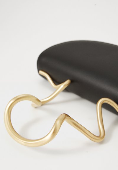Bolsa preta texturizada com uma alça de metal dourado curvada com um design ondulado, destacando materiais contrastantes e um design moderno.