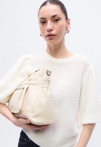 Τσάντα χειρός από μπεζ δέρμα με καμπυλωτό σχήμα, διαθέτει τσέπη με φερμουάρ και λεπτομέρεια με πιέτες. Φοριέται από άτομο ντυμένο με ανοιχτόχρωμο πουλόβερ.