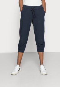 Marineblauwe cropped pants met een drawstring tailleband, zijzakken en elastische manchetten. Gecombineerd met witte sneakers tegen een effen achtergrond.