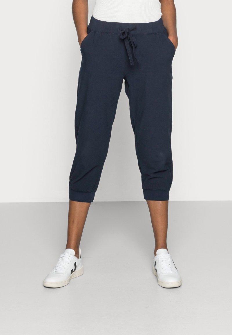 Marineblauwe cropped pants met een drawstring tailleband, zijzakken en elastische manchetten. Gecombineerd met witte sneakers tegen een effen achtergrond.
