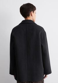 Zwarte wollen jas met een gestructureerd ontwerp, voorzien van een reverskraag, afhangende schouders en een rechte snit. Gladde textuur, geen zichtbare hardware.