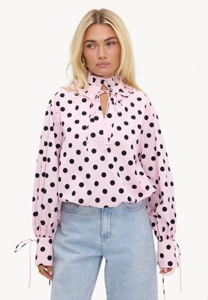 Femme blonde portant un chemisier rose à manches longues avec des pois noirs, col haut à volants, liens aux poignets et au cou, associé à un jean bleu clair.