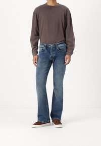 Dlouhý rukáv, tmavě šedá bavlněná košile v kombinaci s modrými džínami. Džíny mají regular fit a mírné vyblednutí. Hnědé tenisky dotvářejí vzhled.