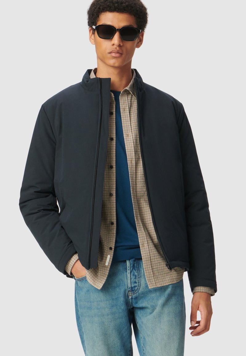 Giacca puffer blu navy con colletto alto, chiusura con zip e vestibilità dritta, indossata sopra una camicia a quadri e una maglietta blu, abbinata a jeans chiari.