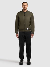 Olivgrüne Bomberjacke mit Frontreißverschluss und gerippten Bündchen, kombiniert mit schwarzen Cargo-Hosen und schwarzen Stiefeln, vor einem neutralen Hintergrund stehend.