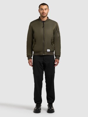 Giubbotto bomber verde oliva con zip frontale e polsini a coste, abbinato a pantaloni cargo neri e stivali neri, in piedi davanti a uno sfondo neutro.