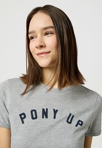 Camiseta gris de tela suave con mangas cortas y un escote redondo. En la parte delantera se muestra un atrevido texto negro "PONY UP".