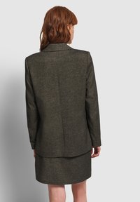 Mujer pelirroja vestida con un traje de falda gris oscuro, de pie con la espalda hacia la cámara frente a un fondo claro gris.