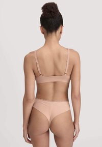 Vrouw gezien van achteren in een bijpassende huidskleurige beha en string ondergoed, tegen een effen lichte achtergrond.