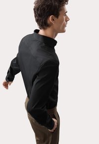 Camicia nera a maniche lunghe con colletto, realizzata in tessuto morbido e con polsini abbottonati, indossata con pantaloni marroni. Design semplice e sartoriale.