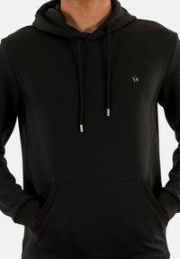 Haut à capuche noir en tissu doux, avec une poche kangourou, des cordons avec des embouts en métal et un petit logo sur la poitrine.