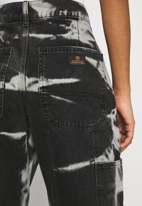 Pantalones cargo de tie-dye en blanco y negro con un tejido de denim texturizado, grandes bolsillos de parche y una etiqueta de marca en la parte trasera.