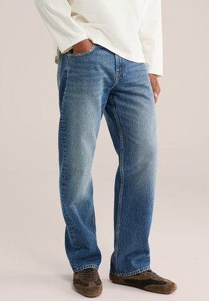 Homme portant un jean droit bleu clair, une chemise blanche à manches longues et des chaussures décontractées marron, debout avec une main dans la poche.
