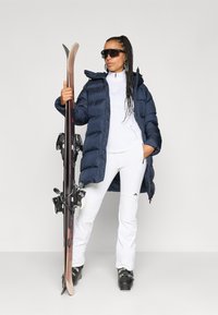 Jachetă puffer albastră, top alb cu fermoar și pantaloni de schi albi. Ținând schiuri cu legături negre, bocanci de schi negri și ochelari de soare. Texturi netede.