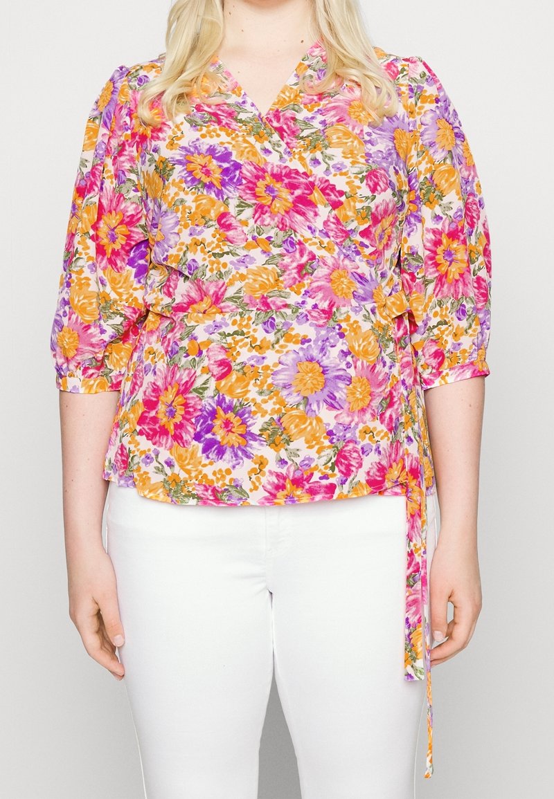 Blusa avvolgente floreale in bianco, con fiori vivaci in rosa, arancione e viola. Maniche corte a sbuffo e dettaglio in vita con nodo. Combinata con pantaloni bianchi.