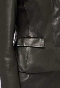 Schwarze Lederjacke mit glatter Oberfläche, ausgestattet mit einer seitlichen Tasche mit Klappe und sichtbaren Nahtdetails an den Ärmeln und am Körper.