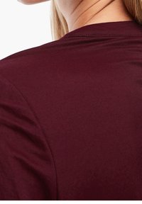 Gros plan sur le haut du dos et le cou d'une personne portant une chemise bordeaux avec des coutures visibles et une texture de tissu lisse.