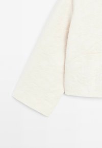 Manche longue en tissu blanc avec une texture jacquard florale délicate et un large détail de manchette sur un fond clair.