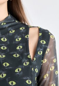 Black and green patterned top featuring eye motifs, with a high neckline and open shoulder design, made from a lightweight fabric.