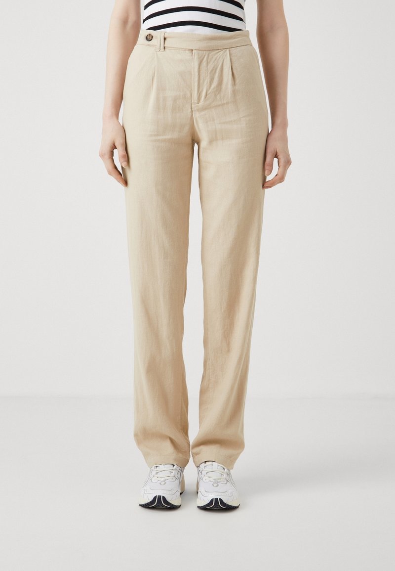 Personne portant un pantalon beige taille haute à jambes droites avec passant de ceinture et un haut sans manches rayé, associé à des baskets blanches.