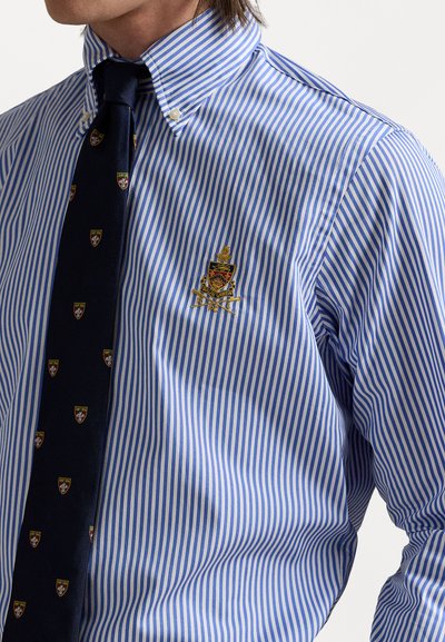 Plava i bijela prugasta košulja na dugmad s ovratnikom i izvezenom emblemu na prsima. Ima tamnu kravatu s uzorkom logotipa.