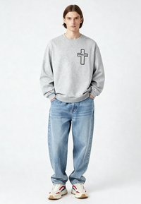 Jeune personne debout, les mains dans les poches, portant un sweat-shirt gris avec une croix et le texte « KARMA KILLS », un jean bleu ample et des baskets blanches.