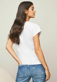 Hvid kortærmet t-shirt med en afslappet pasform, udstyret med en rund halsudskæring og diskrete guldknapdetaljer på skulderen. Bæres med blå jeans.