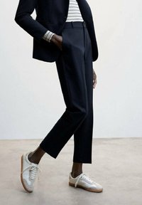 Personne portant un pantalon noir sur mesure, un blazer noir, une chemise à manches longues rayée et des baskets blanches, debout sur un sol neutre.