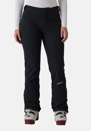 Personne portant un pantalon de ski Spyder noir et des chaussures de ski blanches avec des semelles bordeaux, debout sur un fond clair.