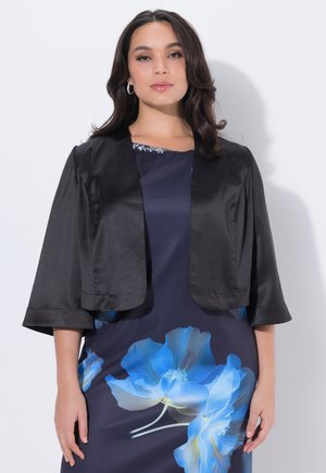 Mujer con una chaqueta corta negra sobre un vestido azul marino con grandes estampados florales azules, de pie frente a un fondo claro y liso.