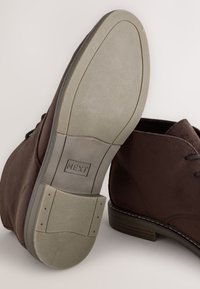 Hnedé semišové členkové topánky s textúrovanou gumovou podrážkou. Majú šnúrky a kontrastnú svetlo šedú podrážku s miernym podpätkom.