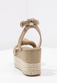 Sandalo wedge in tessuto dorato con cinturino alla caviglia e suola in juta intrecciata, visto da dietro su una superficie bianca.