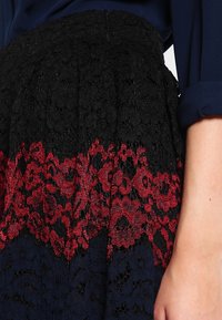 Falda de encaje negro con un patrón floral rojo en el dobladillo, que presenta un diseño texturizado y tejido en capas para mayor dimensión.
