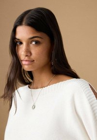 Beli pleteni pulover z širokim izrezom, okrašen z rebrastimi poudarki in izrezanimi detajli. Ogrlica iz srebra z obeskom sonca je na njem.