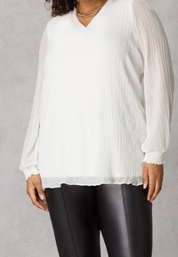 Donna che indossa una blusa bianca a maniche lunghe texturizzata con maniche trasparenti e pantaloni in pelle nera attillati, su uno sfondo neutro.